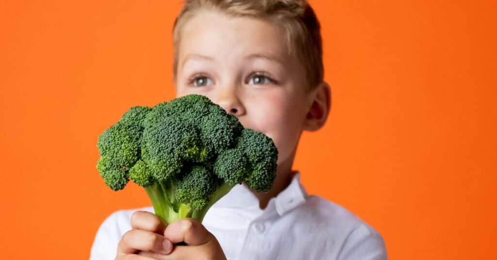 A cheerful young boy holds fresh broccoli, promoting healthy eating against a vibrant orange backdrop.