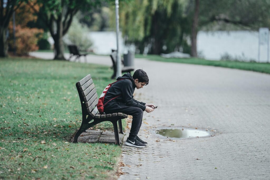 Teen boy sitting on a park bench using his smartphone, surrounded by nature.